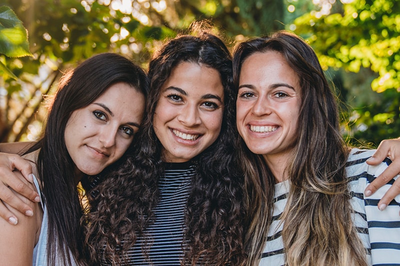 Three smiling friends embracing outdoors in dappled sunlight at Delta Smiles in Antioch and Brentwood, CA